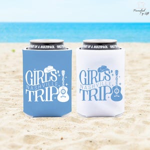 Pu&ograve; includere: Due porta-lattine, uno azzurro e uno bianco, con la scritta "GIRLS NASHVILLE TRIP" e una grafica di chitarra e cappello da cowboy. I porta-lattine sono su una spiaggia sabbiosa con l'oceano sullo sfondo.