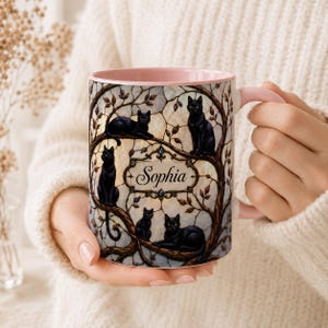 May include: A ceramic mug with a light pink interior and handle, featuring a stained-glass style design of four black cats perched on tree branches. A central decorative frame contains the name Sophia written in a black cursive script font.