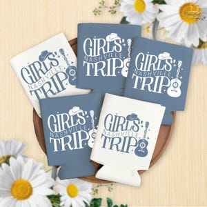 Pu&ograve; includere: Una collezione di portabevande blu e bianchi con la scritta "GIRLS NASHVILLE TRIP 2025". Il design include un cappello da cowboy, una chitarra e delle stelle. I portabevande sono disposti su un vassoio di legno con margherite.