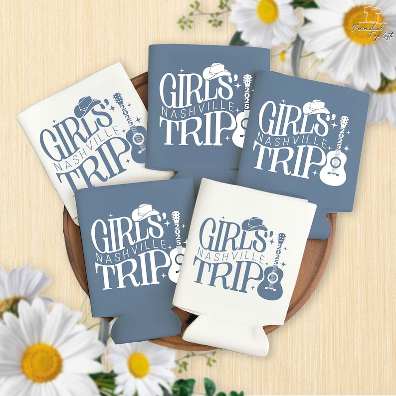 Pu&ograve; includere: Una collezione di portabevande blu e bianchi con la scritta "GIRLS NASHVILLE TRIP 2025". Il design include un cappello da cowboy, una chitarra e delle stelle. I portabevande sono disposti su un vassoio di legno con margherite.