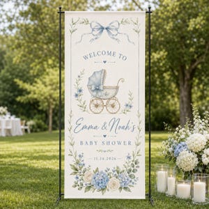 May include: A vertical banner on a black metal stand featuring a blue bow, a vintage baby carriage illustration, and the text "WELCOME TO Emma & Noah's BABY SHOWER 11.26.2026" surrounded by blue and white floral accents on a cream-colored fabric background.