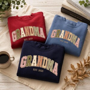 May include: Three crewneck sweatshirts in red, blue, and navy blue. Each features the word GRANDMA in arched, multi-colored gingham plaid letters with the text EST. 2025 printed underneath in a simple, clean font on the front of the garment.