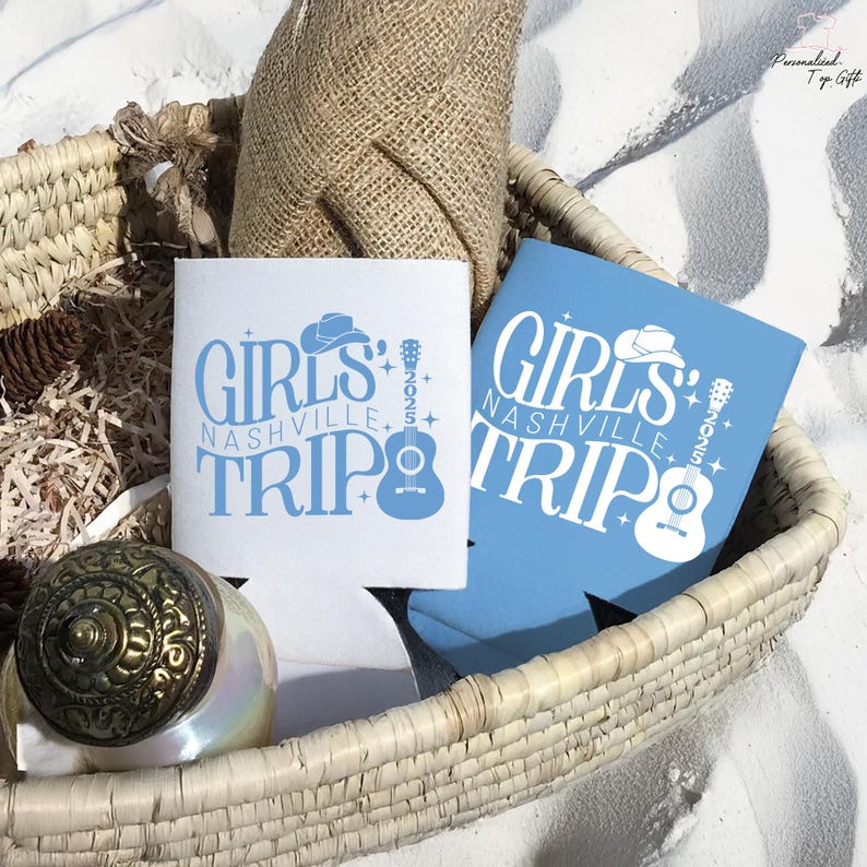 Pu&ograve; includere: Due isolanti per bevande, uno bianco e uno azzurro chiaro, con la scritta "GIRLS NASHVILLE TRIP 2025". Il design include un cappello da cowboy, una chitarra e stelle. Gli isolanti sono in un cestino intrecciato.