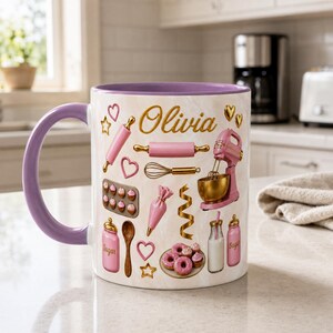 May include: A ceramic mug with a light purple handle and interior, featuring a marble-patterned exterior decorated with gold and pink baking-themed illustrations, including a stand mixer, rolling pin, whisk, and the name Olivia in gold script.