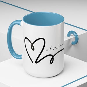 May include: A white ceramic mug with a light blue interior and handle. The exterior features a black line drawing of a heart shape followed by Morse code symbols. The design is printed on the front of the mug, which has a smooth, cylindrical body.