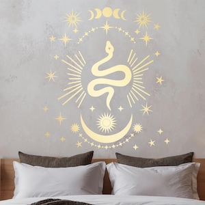 Könnte beinhalten: Ein weißes Bett mit einer braunen Strickdecke. Die Wand hinter dem Bett hat ein goldenes Schlangen- und Mond-Dekor mit Sternen.