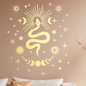 Könnte beinhalten: Goldene Wanddekoration mit einer Schlange, über der ein Sonnenstrahl und unter der ein Halbmond zu sehen sind. Die Dekoration ist von Sternen und Monden umgeben. Die Dekoration befindet sich an einer hellbraunen Wand mit einem weißen Sofa und einer Vase mit getrocknetem Gras im Vordergrund.