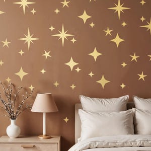 Op de afbeelding: Een slaapkamermuur is versierd met gouden sterstickers van verschillende formaten. Een nachtkastje met een lamp en een vaas met bloemen staat naast een bed met wit beddengoed. De muur is geschilderd in een warme bruine kleur.