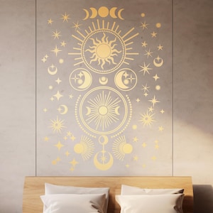 Peut inclure: Un autocollant mural céleste doré avec un motif de soleil, de lune et d'étoiles. L'autocollant est appliqué sur un mur gris clair au-dessus d'une tête de lit en bois et d'une literie blanche.