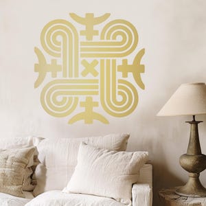 Peut inclure: Un autocollant mural doré avec un motif géométrique abstrait. Le motif est composé de lignes et de formes interconnectées, créant un motif symétrique. L'autocollant est monté sur un mur de couleur claire au-dessus d'un canapé blanc avec des coussins et une lampe.