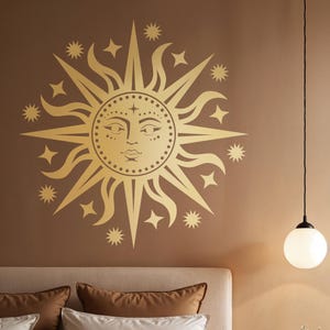 Puede incluir: Un vinilo decorativo dorado de sol y estrellas con una cara, fijado a una pared marrón. El diseño del sol está rodeado de estrellas. Una lámpara colgante de globo blanco cuelga del techo. La cama tiene almohadas marrones y blancas.