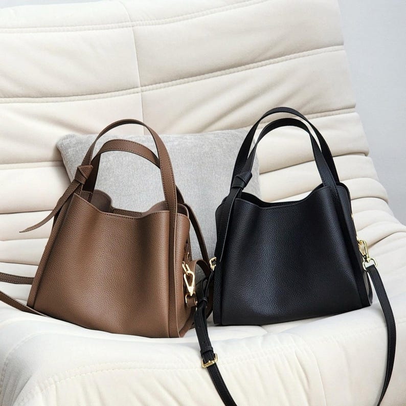 May include: Two leather tote bags, one brown and one black, with a gold clasp on the brown bag. The bags are sitting on a white couch.
