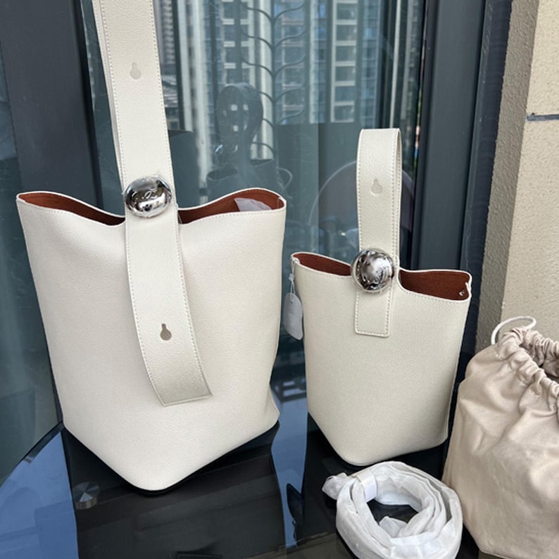 May include: Two white leather bucket bags with silver hardware. The larger bag has a long strap and the smaller bag has a short handle. Both bags have a round silver buckle on the front.