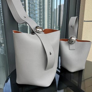 May include: Two gray leather bucket bags with silver hardware. The larger bag has a long strap and a top handle. The smaller bag has a top handle only.