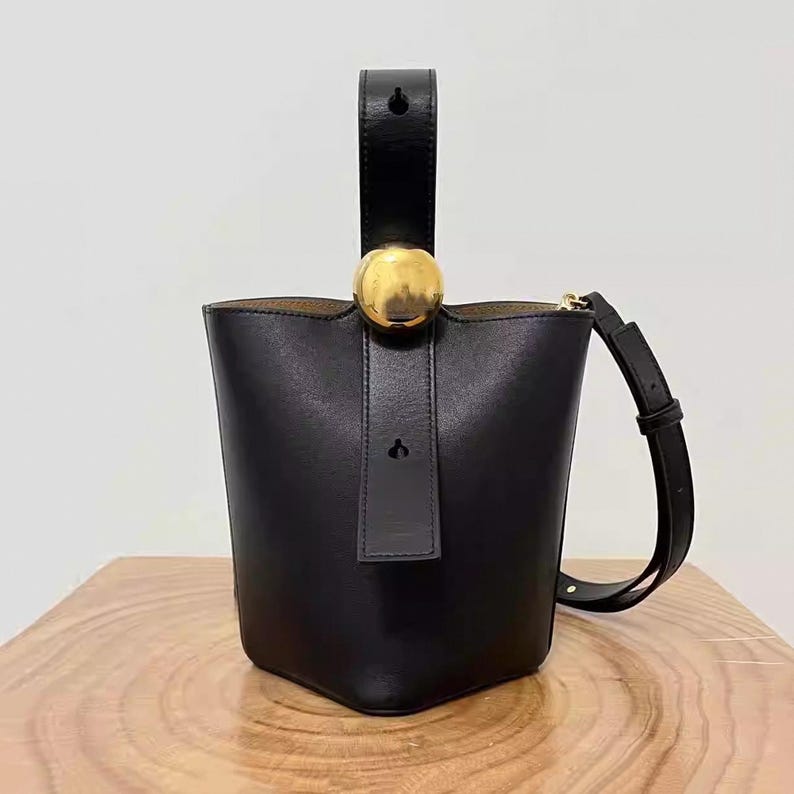 May include: Black leather bucket bag with a gold top handle and a gold sphere accent. The bag has a long strap that can be worn on the shoulder or crossbody.