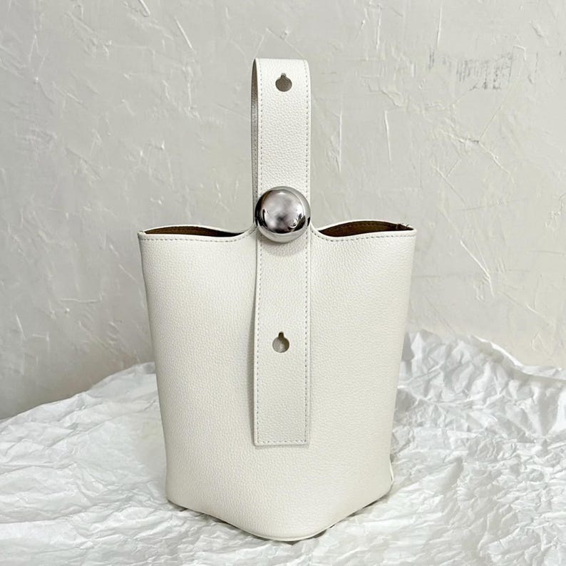 May include: A white leather bucket bag with a silver top handle and a silver button closure.
