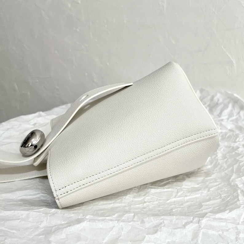May include: A white leather bucket bag with a silver metal accent. The bag has a top handle and a detachable strap.
