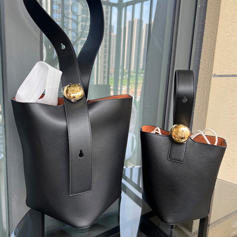 May include: Two black leather bucket bags with gold hardware. The larger bag has a top handle and a long strap. The smaller bag has a top handle.