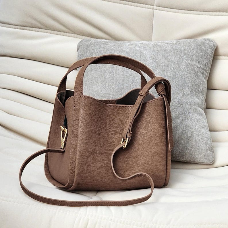 May include: A brown leather tote bag with a top handle and a long shoulder strap. The bag has a knotted detail on the strap and a gold-toned buckle.