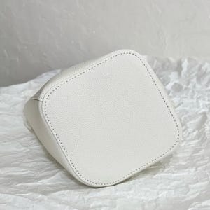 May include: White leather bucket bag with a stitched design. The bag is made of a textured material and has a smooth, clean finish.