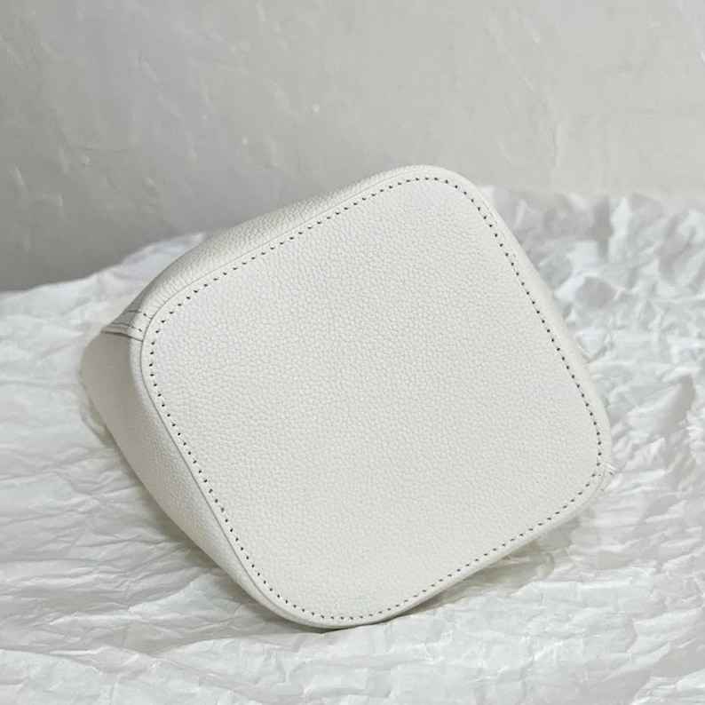 May include: White leather bucket bag with a stitched design. The bag is made of a textured material and has a smooth, clean finish.