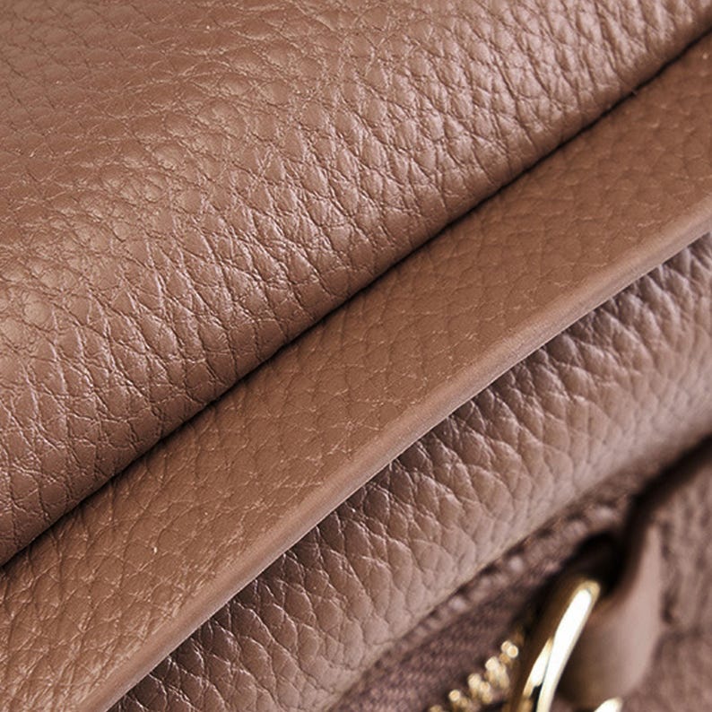 May include: Close-up of a brown leather bag with a zipper detail. The leather has a textured, pebbled surface.
