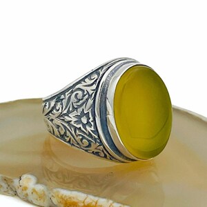 Könnte beinhalten: Ein silberner Ring mit einem ovalen, gelben Edelstein. Der Ring hat ein verziertes, graviertes Design am Band. Der Edelstein ist in einer silbernen Lünette gefasst. Der Ring wird auf einer hellen, gemusterten Oberfläche präsentiert.