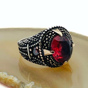 Könnte beinhalten: Ein silberner Ring mit einem großen, ovalen, tiefroten Edelstein. Der Ring hat ein verziertes, strukturiertes Band mit kleinen, dunklen Akzenten und goldfarbenen Details. Der Edelstein ist der Blickfang und fängt das Licht ein.