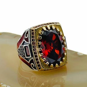 May include: A silver ring with a large, rectangular, deep red gemstone. The ring has a gold-coloured bezel and accents, with smaller red stones around the main stone. The band features ornate detailing in black and red.