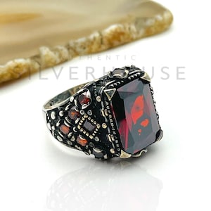 Könnte beinhalten: Ein silberner Ring mit einem großen, rechteckigen, roten Edelstein als Mittelpunkt. Der Ring hat aufwendige Details mit kleineren roten Steinen und einem dunklen, oxidierten Finish. Das Design ist verziert und im Vintage- oder Antik-Stil gehalten.