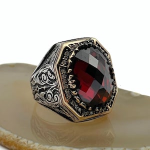 Peut inclure: Bague en argent ornée d'une pierre rouge foncé facettée de forme octogonale. La bague est dotée d'une lunette dorée et ornée de petites pierres claires. La bande présente un motif tourbillonnant orné.