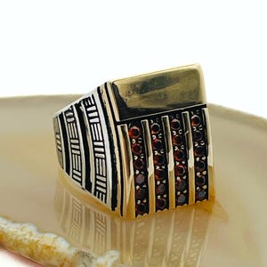 Peut inclure: Une bague en argent et or avec un dessus rectangulaire en or et un design détaillé. La bague présente un motif de lignes verticales et de petites pierres précieuses rouges. La bague est présentée sur une surface claire et à motifs.