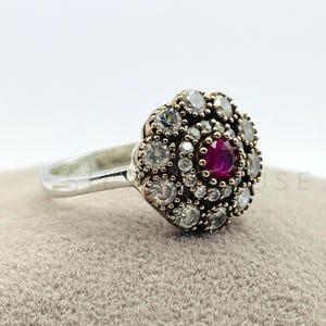 Puede incluir: Un anillo de plata con diseño floral. El centro presenta una gema redonda de color rojo intenso, rodeada de piedras claras más pequeñas. El anillo se encuentra sobre una superficie gris, mostrando los detalles.