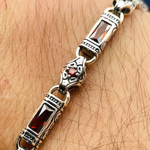 May include: Silver bracelet with rectangular red gemstones and ornate detailing. The bracelet features a series of connected links, with each link showcasing a central red stone. The design incorporates intricate patterns and small red accents.