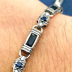 Peut inclure: Bracelet en argent avec des détails ornés et des pierres précieuses rectangulaires et rondes bleu foncé. Le bracelet est en métal brillant et présente un design complexe.