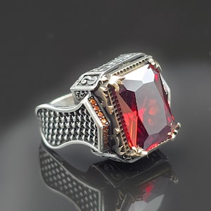 Puede incluir: Un anillo de plata con una gran gema roja rectangular. El anillo tiene detalles intrincados en la banda y alrededor de la gema, con detalles dorados. La gema facetada captura la luz y refleja un color rojo intenso.
