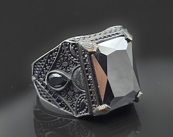 May include: A silver ring with a large, rectangular black gemstone. The ring features intricate, detailed patterns on the band and smaller black stones. The gemstone is the focal point, reflecting light.