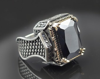 May include: A silver ring with a large, rectangular black gemstone. The ring has intricate detailing on the band and around the stone, with gold accents. The ring is displayed on a reflective surface.