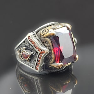 Peut inclure: Bague en argent ornée d'une grande pierre rouge foncé de forme rectangulaire. La bague présente des détails complexes, notamment de petites pierres rouges et des accents dorés. Le design est orné, sur fond sombre.