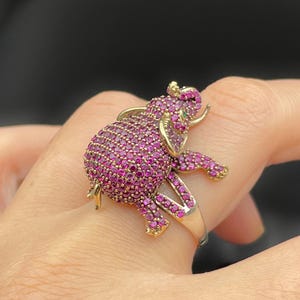Peut inclure: Une bague dorée en forme d'éléphant, ornée de nombreuses petites pierres précieuses roses vives. Le corps de l'éléphant est entièrement recouvert de pierres roses, avec des accents dorés sur les oreilles, les défenses et les pattes. Une petite pierre verte est visible sur l'œil.