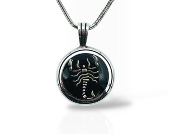 May include: A silver necklace with a circular pendant. The pendant has a black background with a silver scorpion design. The chain is a fine, textured style, and the pendant is suspended from a small bail. The pendant's diameter is approximately 2.5 cm.