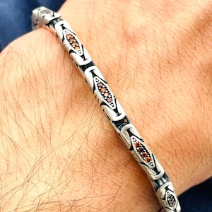 May include: Silver bracelet with an ornate design. The bracelet features a series of interconnected links, each with a detailed pattern. Small, reddish-brown gemstones are set within the links, adding a touch of color and sparkle to the jewelry.