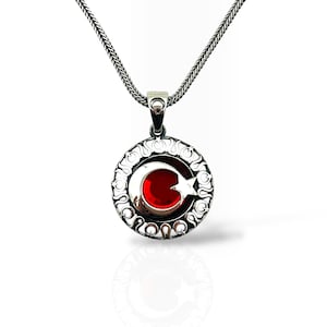May include: A silver pendant necklace. The circular pendant has a red centre with a crescent moon and star design, framed by an ornate border. The chain is a textured, silver-coloured link.