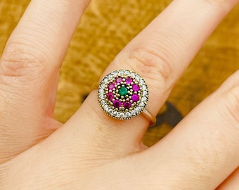 Può includere: Un anello d'oro con un centro rotondo che presenta una pietra verde circondata da pietre rosa e un bordo di pietre bianche.