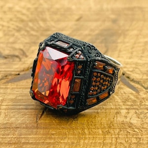 May include: A black ring with a large, red, square-shaped gemstone. The ring has intricate details and smaller red gemstones around the main stone.