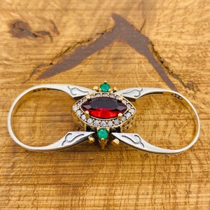 Könnte beinhalten: Ein Silber- und Goldring mit einem roten Edelstein, der von kleineren weißen Edelsteinen umgeben ist. Der Ring hat zwei Bänder, die durch ein dekoratives Design verbunden sind.