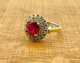 Könnte beinhalten: Ein goldener Ring mit einem großen roten Edelstein, der von kleineren weißen Edelsteinen umgeben ist.
