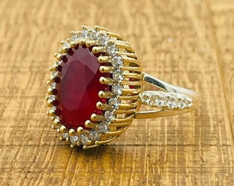 Könnte beinhalten: Ein goldener Ring mit einem großen roten ovalen Edelstein, der von kleineren weißen Edelsteinen umgeben ist. Der Ring hat eine silberne Band.