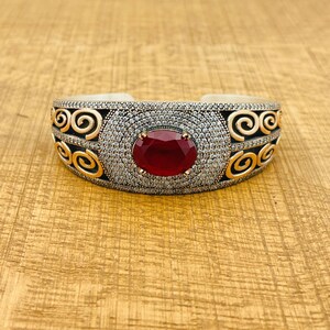 Peut inclure: Un bracelet en argent avec un motif en spirale doré et une pierre rouge. Le bracelet est serti de pierres claires.