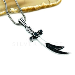 May include: A silver-toned pendant necklace featuring a stylized dagger design. The dagger has a curved blade with black accents and a decorative handle. The necklace chain is a textured, linked design.